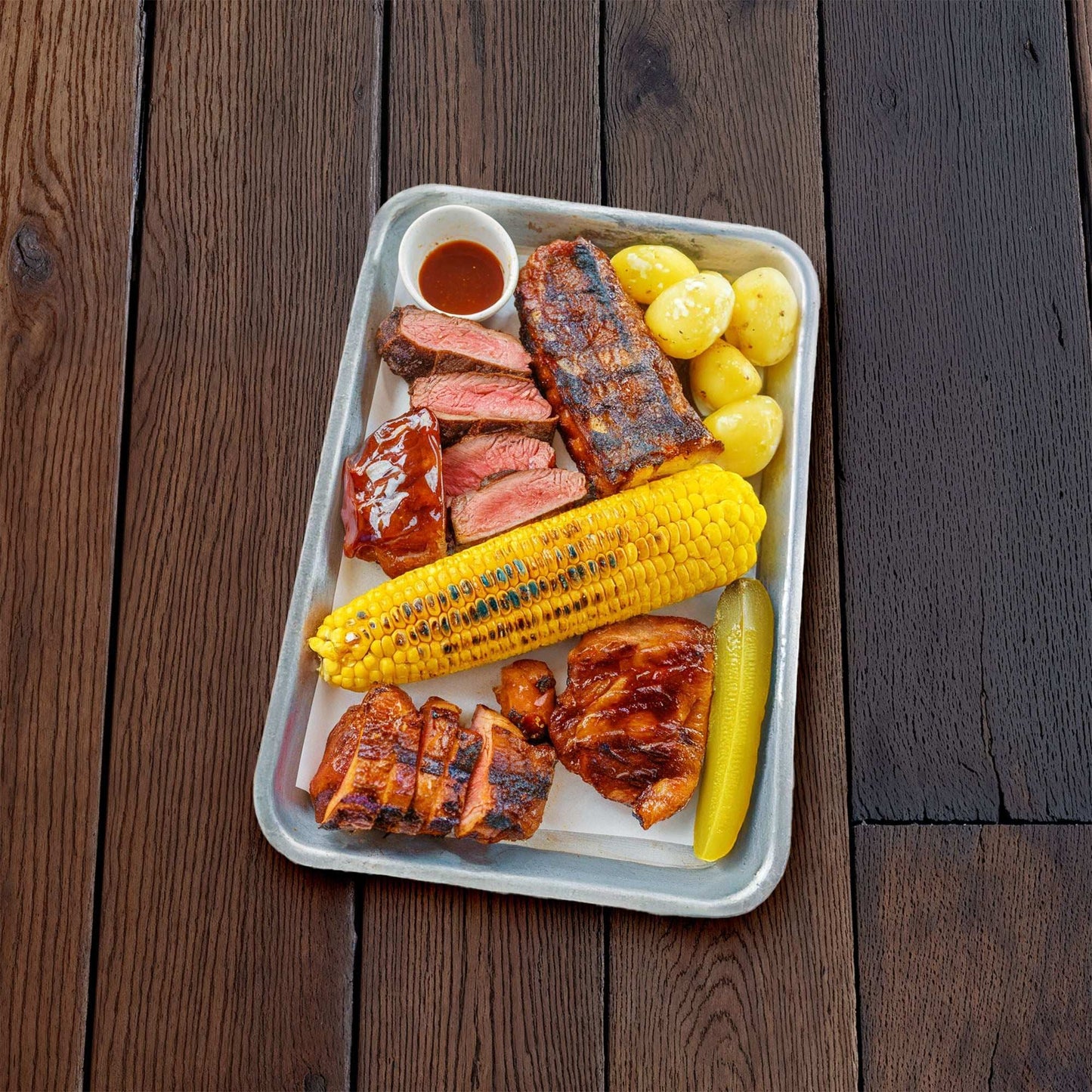 Dark oak saloon floor backdrop with textured wood planks, showcasing a meal of grilled meats and vegetables on a tray.