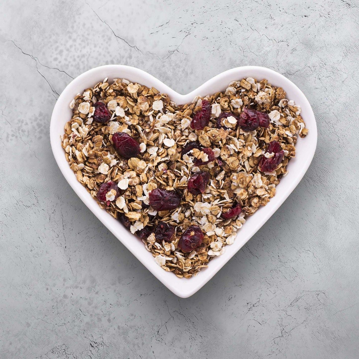 Neutral gray stone texture backdrop with heart-shaped bowl of oats and cranberries.
